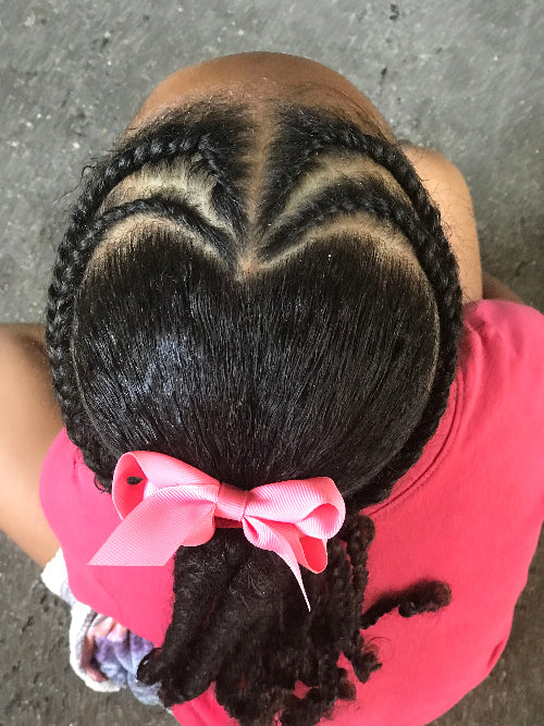 Coiffure d’enfant afro avec tracé en forme de cœur et tresses collées, accessoirisée avec un nœud rose.