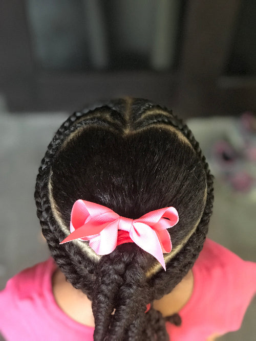 Vue du dessus d’une coiffure pour enfant avec tresses collées en forme de cœur et nœud rose, sur cheveux texturés.