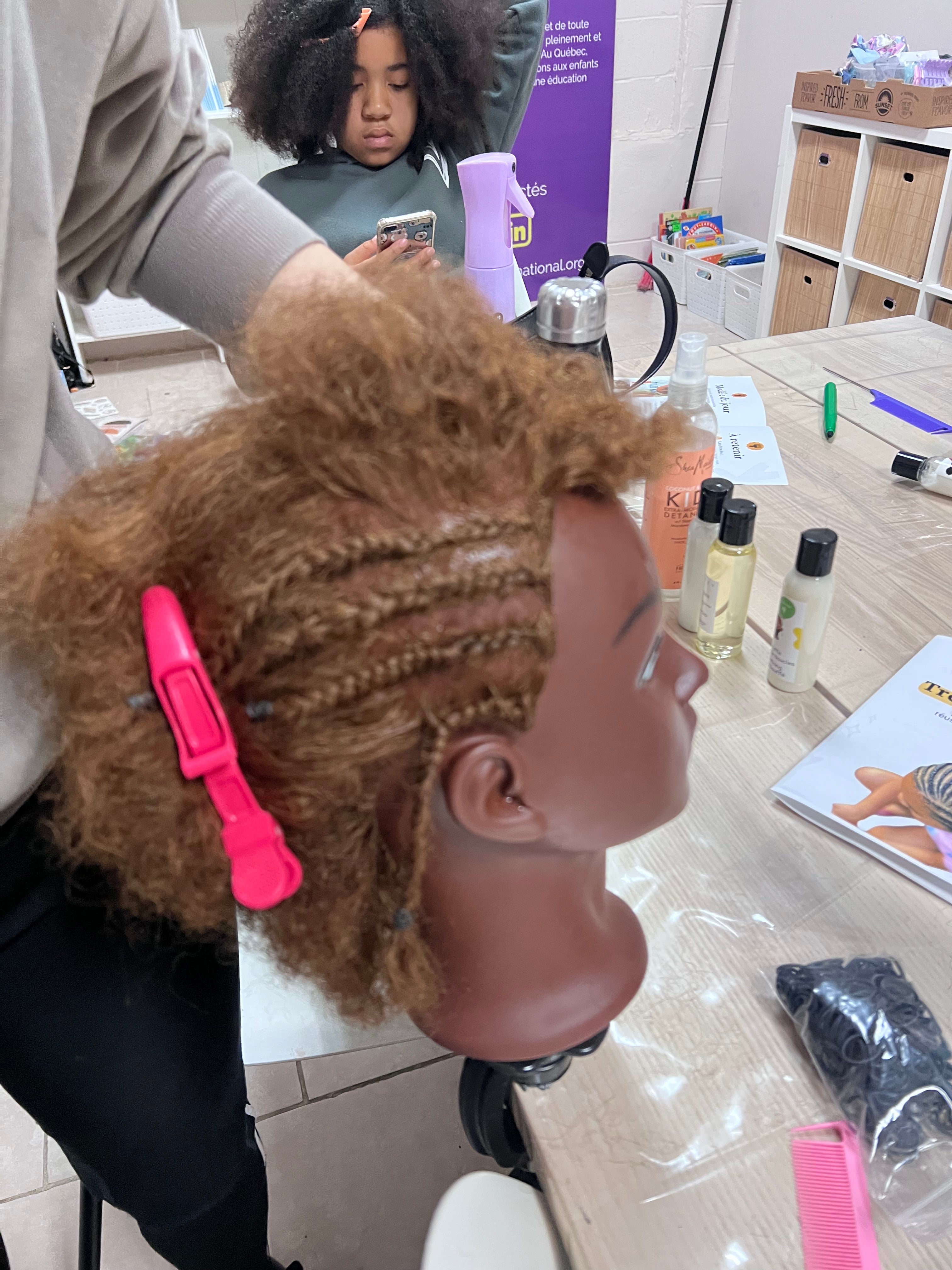 Vue de profil d’une tête à coiffer aux cheveux cuivrés avec plusieurs tresses fines et une pince rose.
