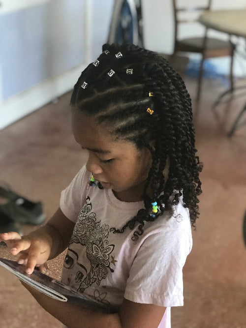 Petite fille avec coiffure twist décorée de perles et bijoux, en pleine séance de coiffage dans un tuto vidéo.